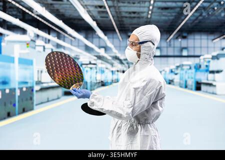 Man in bunny suit evaluating silicon wafer in brightly lit manufacturing plant cleanroom. Engineer checking semiconductor substrate nanometer circuit in modern foundry facility Stock Photo