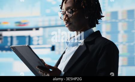 Manager using tablet to compare company metrics on digital videowall before making decisions. Entrepreneur analyzing key performance indicators using device app to optimize profitability, camera B Stock Photo