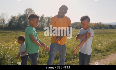 Father enjoying a sunny day in the countryside with his three sons, helping his older twin boys apply sunscreen correctly. Family time concept. Stock Photo