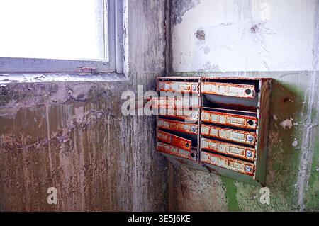 Ukraine, Pripyat : Set of old metal mailboxes heavily rusted and open mounted on peeling painted wall next to window in the entrance hall of an abando Stock Photo