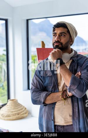 Asian man in 30s standing in personal studio near windows with easel, red canvas, headphones Stock Photo