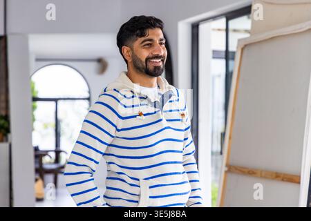 Bearded man wearing white and blue striped hoodie smiling near canvas on easel in home studio Stock Photo