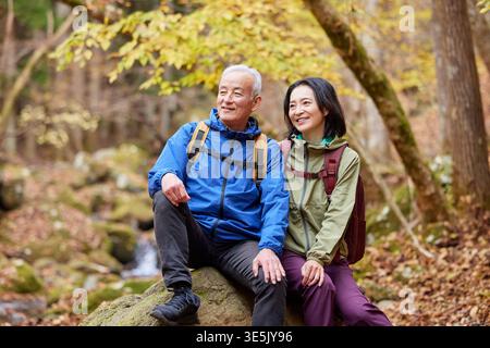 Smiling Japanese senior couple resting on rock during autumn forest hike Stock Photo