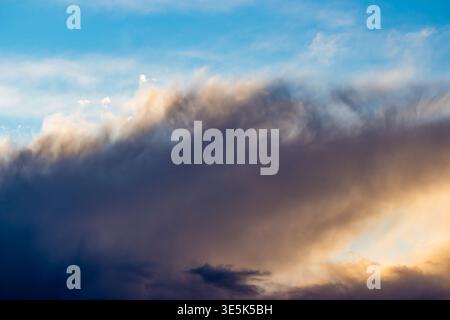 Dramatic, colorful cloudscape with dark storm clouds contrasting golden illuminated edges against a serene blue sky. Dynamic weather pattern, ethereal Stock Photo
