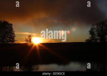 Radiant sunset over a tranquil lake, sunbeams bursting through dark clouds. Silhouetted trees frame the vibrant sky and water reflections with atmosph Stock Photo