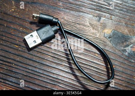 Damaged black USB-A to Micro-USB cable reveals frayed wires on a dark rustic wooden surface. Symbolizing repair, connectivity issues, and obsolete tec Stock Photo