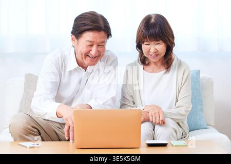 Smiling Japanese senior couple enjoying online communication and information gathering on a laptop in their living room Stock Photo