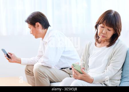 Japanese senior couple using smartphones on a sofa in a living room ignoring each other Stock Photo