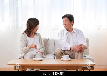 Smiling Japanese senior couple relaxing and talking with coffee cups on sofa in bright living room Stock Photo
