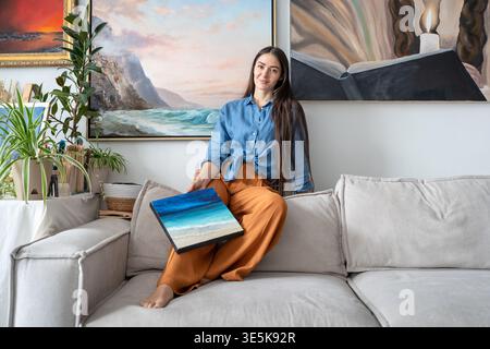 Happy talented woman painter posing at camera with artwork sitting on couch at home loft gallery. Stock Photo
