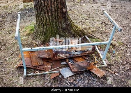 Scrap metal heap, featuring rusty pieces and a light blue frame, discarded at the base of a large tree trunk in a barren outdoor setting Stock Photo