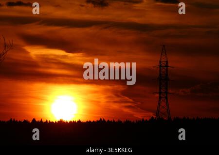 Stunning sunset glows behind a tall electric pylon and a dark forest silhouette, creating a dramatic, vivid skyline Stock Photo