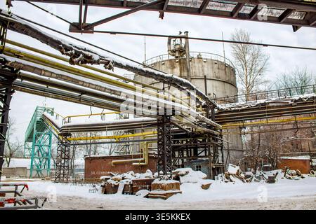 Snow-covered pipes and rusty industrial frameworks crisscross against a wintry sky. A huge concrete silo anchors the grimy, cold factory yard scene Stock Photo