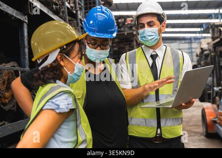 Diverse workers in face masks with laptop discussing inventory in auto parts warehouse Stock Photo
