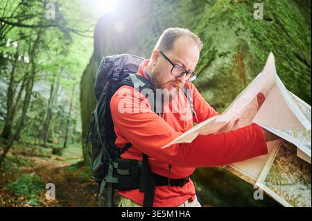 Bearded tourist man looks puzzled, studies map while stands in front of large rock in dense forest. Traveler with grey backpack and glasses, trying to figure out his direction. Stock Photo