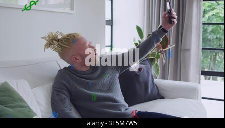Holding smartphone, man in gray sweater leaning on white sofa with throw-pillows near black-windows Stock Photo