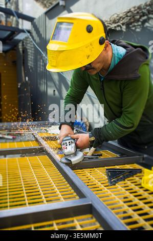 A worker cuts metal with a tool on a construction site. Sparks fly as he focuses on his task. The bright yellow grid beneath him adds color to the sce Stock Photo