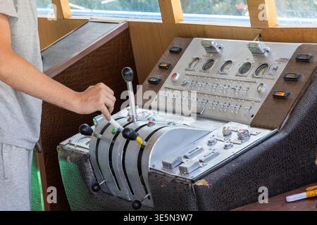 Adult man focuses on manipulating control panel with levers and switches. Large window reveals greenery outside, enhancing operational mood. Stock Photo