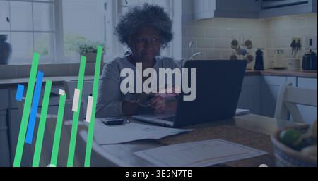 Sitting senior woman wearing grey top with reading glasses, checking papers using laptop in kitchen Stock Photo