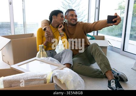 Smiling Diverse couple sitting on floor in modern living room, with smartphone and moving boxes Stock Photo