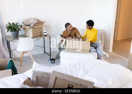 Sorting Diverse couple unpacking bubble wrap on ottoman in bedroom, with cardboard boxes Stock Photo