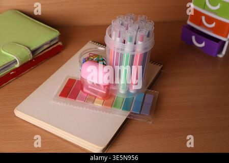 Bright colored markers and a pink stapler sit on a notepad on a wooden desk. Nearby, there are paper clips and small organizers holding other supplies Stock Photo
