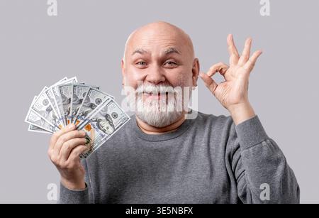 Senior man holding dollars banknotes doing ok sign fingers, smiling friendly gesturing excellent symbol isolated on gray background. Man holding Stock Photo