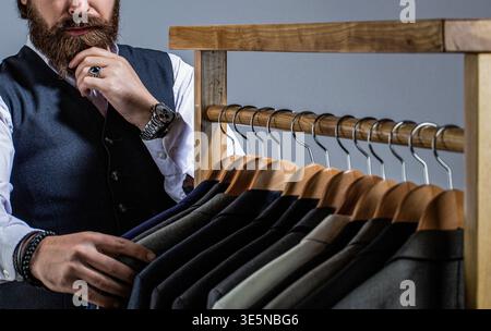 Male suits hanging in a row. Stylish man suit. Man suit, tailor in his workshop. Handsome bearded fashion man in classical costume suit Stock Photo