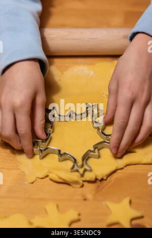 Child's hands press a festive metal cookie cutter into rolled yellow shortcrust pastry on a wooden surface. Rolling pin visible. Christmas home baking Stock Photo
