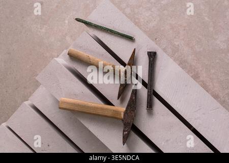 Two rusty stone carving hammers with wooden handles, two chisels and a green pencil arranged on grey stone slabs. Overhead flat lay, authentic artisan Stock Photo