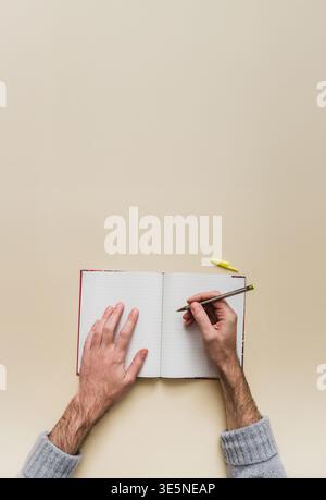 Male hands writing with pencil in open lined notebook from top view on uniform beige background. Left hand holds page, large white area for text. Crea Stock Photo