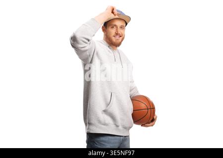Young man in a gray hoodie holding a basketball and tipping his cap isolated on white background Stock Photo