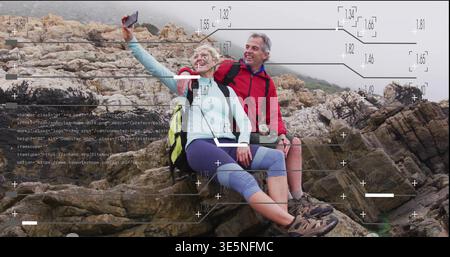 Smiling couple wearing hiking gear sitting on rocky cliff taking selfie with phone, packs, overlays Stock Photo