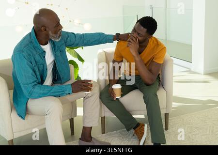 Sitting African American father and son holding paper coffee cups in living room, with globe lights Stock Photo