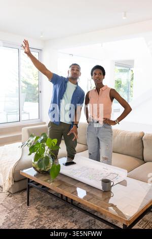 Gesturing couple examining blueprint in modern living room, with coffee mug smartphone and plant Stock Photo