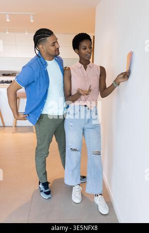 Diverse couple selecting paint color swatch cards on blank white wall in modern kitchen space Stock Photo