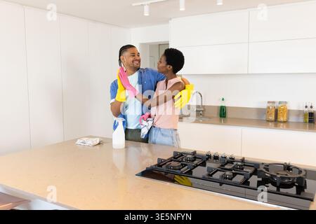 Hugging diverse couple wearing gloves cleaning kitchen counter in bright kitchen, with spray bottle Stock Photo
