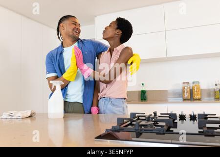 Laughing Diverse couple wearing rubber gloves cleaning countertop in kitchen, with spray bottle Stock Photo