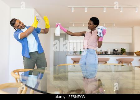 Laughing Diverse couple wearing gloves playfully cleaning in open kitchen, with sprayer and cloths Stock Photo