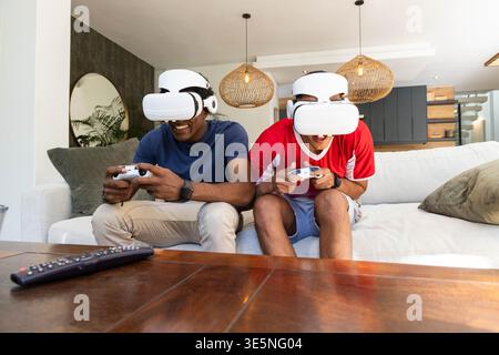 Playing Diverse male friends using VR headsets on sofa in living room, with gaming controllers Stock Photo