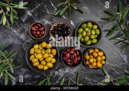 Green and black olives in a bowls and olive oil on dark background top view. Olive oil and variety of green and black olives in bowls with olive leaves all around taken from above. Stock Photo