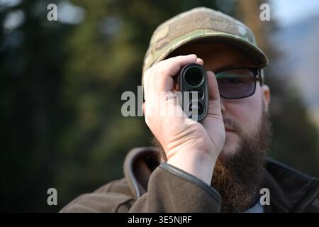A bearded hunter wearing a camouflage cap and glasses looks through a handheld rangefinder in a forest setting, concentrating on distance and scouting while dressed for outdoor hunting and recreation. Stock Photo