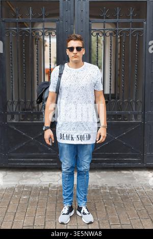 Stylish handsome fashion hipster man in trendy casual clothes with glasses with backpack on the street Stock Photo