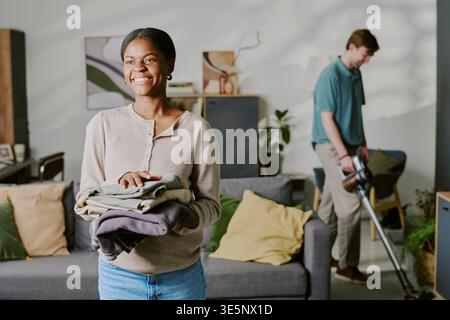 Black Woman Holding Folded Laundry While Man Vacuuming at Home Stock Photo