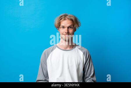 Young man with blond messy hair smiling confidently, featuring a casual raglan shirt and engaging expression on a vibrant blue background Stock Photo