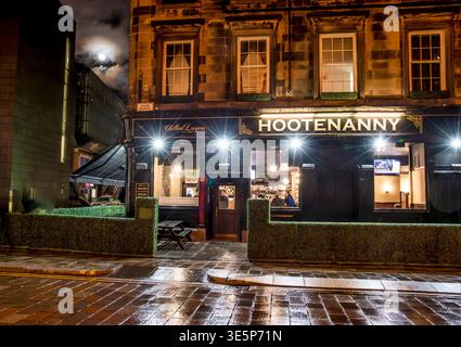 Nighttime exterior of the Hootenanny pub showing warm interior lights, patrons at the bar, wet pavement reflections and atmospheric signage on a histo Stock Photo