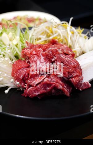Fresh raw beef bulgogi with assorted vegetables including green onions, bean sprouts, and enoki mushrooms on a black plate Stock Photo