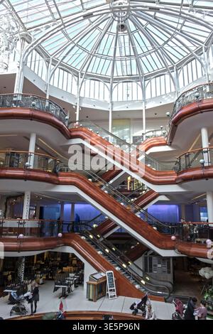 Interior view of Princes Square shopping centre showing intersecting wooden escalators beneath a bright glass dome atrium, with shoppers and multiple Stock Photo
