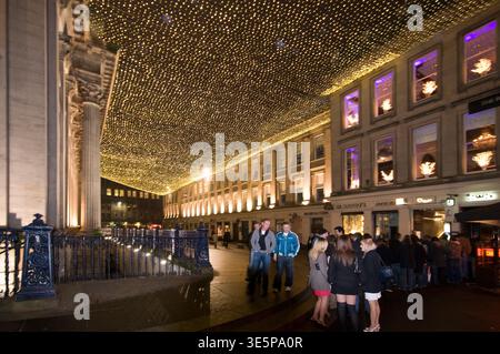 Crowd of friends and visitors gathered on a city street beneath a glowing canopy of decorative lights outside a gallery, creating a festive, lively ni Stock Photo