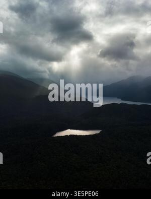 Aerial view of sun rays piercing through stormy clouds over the rugged, shadowed landscape, illuminating a small lake near Manapouri Lake, Manapouri, Stock Photo